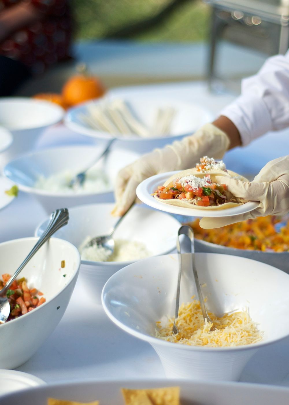 Gloved catering staff plating food at an outdoor buffet with bowls of fresh toppings